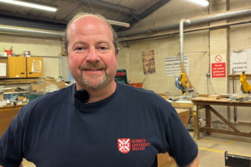 Male member of staff stands in a workshop space with woodworking tools and benches visible in the background.